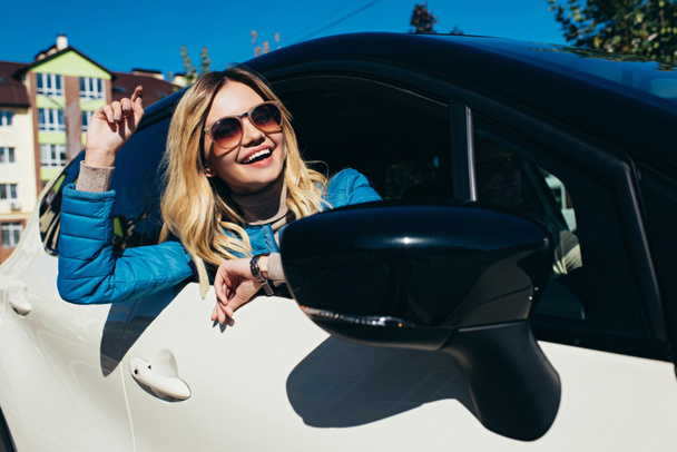 Portret van vrolijke jonge vrouw in zonnebril leunend uit auto op straat - Foto, afbeelding