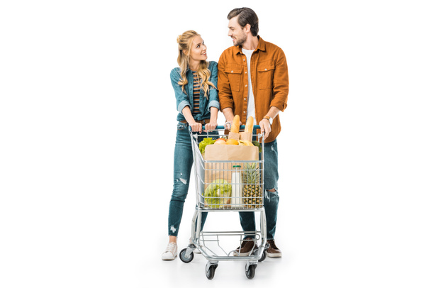 happy couple carrying shopping trolley with products and looking at each other isolated on white - 写真・画像