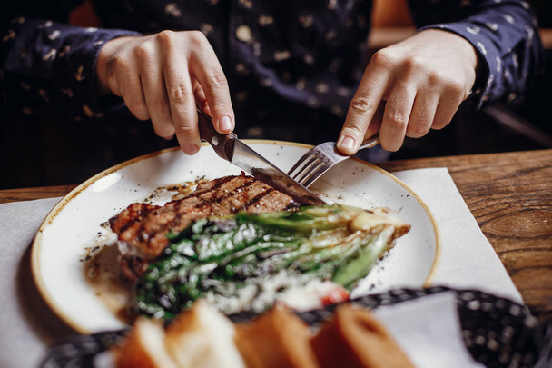 Hands holding fork and knife and eating delicious juicy steak with grilled cabbage,tomatoes and cheese on table at cafe in city street. Man tasting bbq with vegetables in restaurant - 写真・画像
