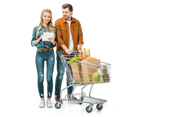 atractiva mujer sonriente revisando la lista de compras en la tableta digital mientras su novio de pie cerca con el carro lleno de bolsas de papel con productos aislados en blanco
 - Foto, Imagen