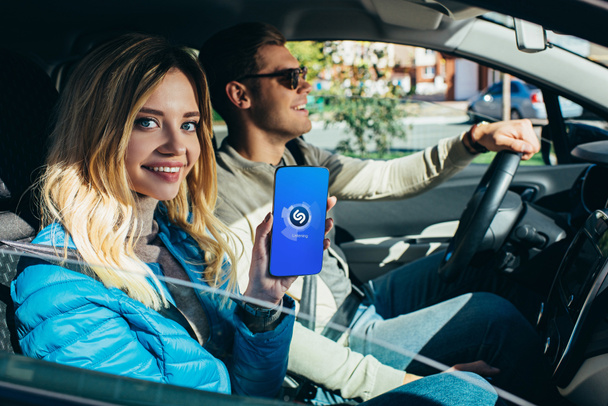 smiling woman showing smartphone with shazam logo on screen while husband driving car - Zdjęcie, obraz
