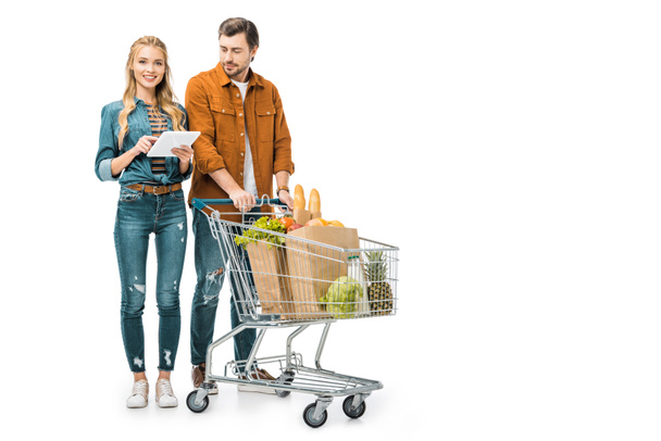 young woman checking shopping list on digital tablet while her boyfriend standing near with trolley full of paper bags with products isolated on white - Photo, Image