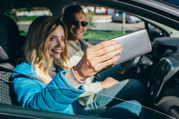 χαμογελαστό γυναίκα λήψη selfie μαζί με το φίλο της στο αυτοκίνητο κατά τη διάρκεια ταξιδιού - Φωτογραφία, εικόνα