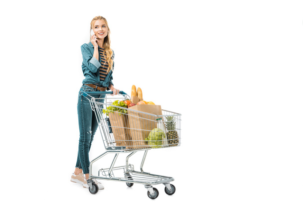 krásná žena mluví o smartphone a nesoucí nákupní vozík s produkty izolované na bílém - Fotografie, Obrázek