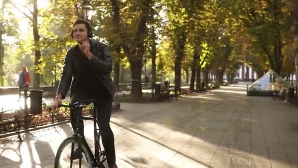 Young caucasian man with headphones riding bicycle in the city park on the trekking black bike. Riding and feeling good while listening to the favourite music. Sun shines on the background - Video