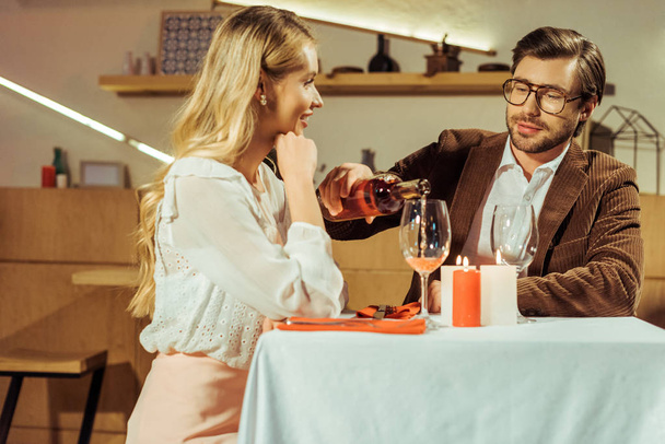man in jacket pouring wine into glass to beautiful girlfriend at table in restaurant  - Photo, Image
