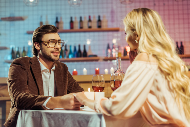 若いカップルは手を繋いでいる笑顔とキャンドルとレストランでワイングラスを持つテーブルでロマンチックな夕食  - 写真・画像