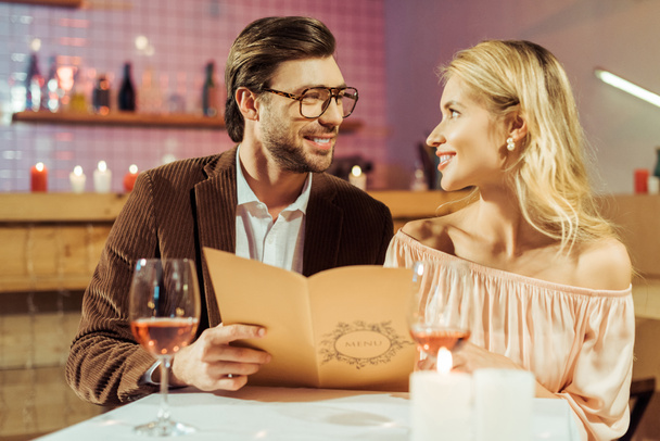 レストランでロマンチックな夕食時にお互いを見てメニューと笑顔のカップル  - 写真・画像