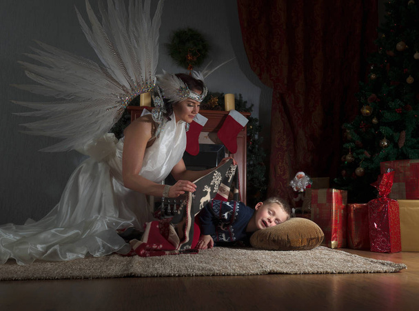Niño dormido bajo el árbol de Navidad y ángel guardián cubriéndolo con una manta. Angel en vestido blanco con plumas naturales
. - Foto, imagen
