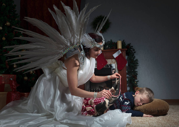 Niño dormido bajo el árbol de Navidad y ángel guardián cubriéndolo con una manta. Angel en vestido blanco con plumas naturales
. - Foto, Imagen