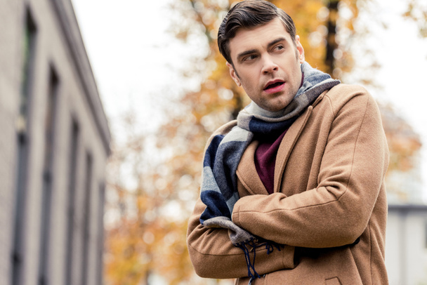 bottom view of handsome man in coat freezing on autumnal street - Fotografie, Obrázek