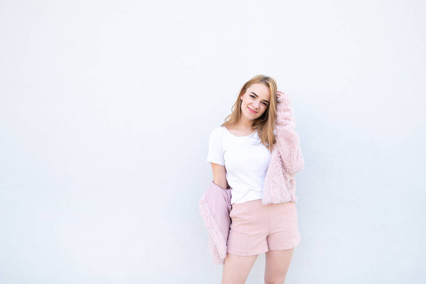 Sonriendo hermosa adolescente en pantalones cortos de color rosa y un abrigo de pie sobre un fondo blanco, mirando a la cámara y sonriendo. Espacio de copia
 - Foto, Imagen