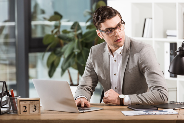 handsome confident businessman sitting at workplace and looking away in modern office - Foto, Imagen