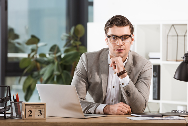 handsome confident businessman sitting at workplace and looking at camera in modern office - Photo, Image