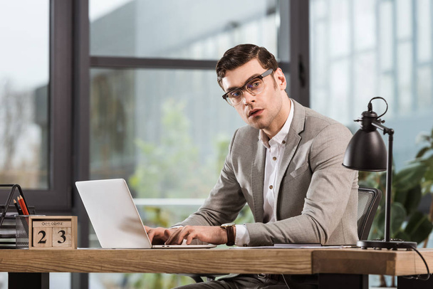 handsome businessman working with laptop and looking away at office - Foto, imagen