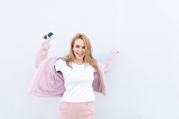 Trendy meisje in een wit T-shirt en een roze jas met een smartphone in haar handen en een koptelefoon, kijkt naar de camera en verheugt zich op een witte achtergrond. Gelukkig meisje op de achtergrond van een witte muur. - Foto, afbeelding