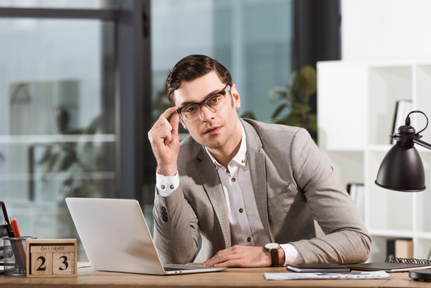 handsome businessman with eyeglasses sitting at workplace in office and looking at camera - Фото, изображение