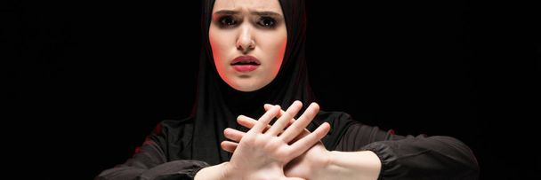 Portrait of beautiful desperate scared frightened young muslim woman wearing black hijab showing stop sign on black background - Photo, Image