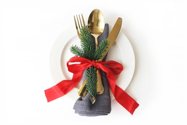 Traditional Christmas table place setting. Golden cutlery, linen napkin, green spruce branches, plate and red ribbon decoration isolated on white background. Holidays background. Flat lay, top view - Φωτογραφία, εικόνα