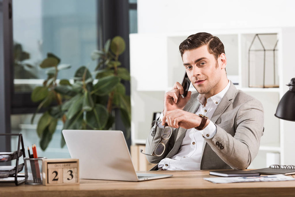 handsome businessman talking by phone at workplace in office and looking at camera - Foto, Bild