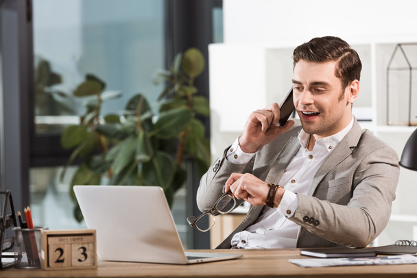 handsome happy businessman talking by phone at workplace in office and looking at laptop screen - Foto, Bild