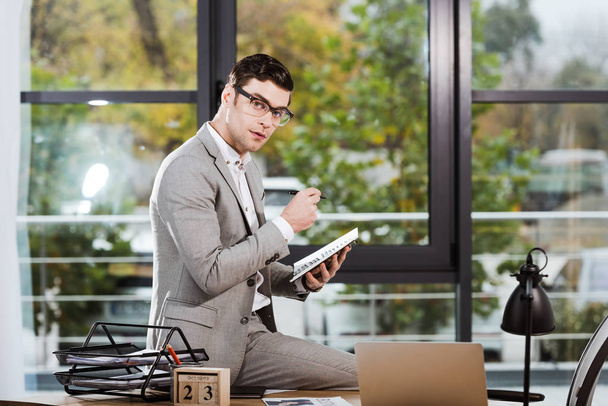 pohledný podnikatel s notebookem sedět na pracovišti v kanceláři a při pohledu na fotoaparát - Fotografie, Obrázek