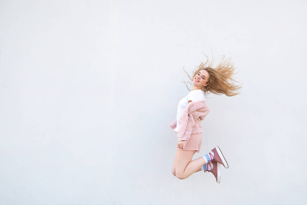 Young blonde girl in pink clothes jumping against the background of a white wall and smiling. Attractive happy girl levitation on the background of a white wall. Copyspace - Foto, Bild