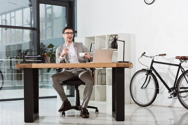 handsome happy businessman with cup of coffee sitting at workplace in modern office - Φωτογραφία, εικόνα