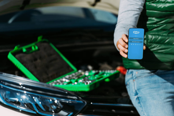cropped shot of man holding smartphone with skype app while sitting on broken car with toolbox - Zdjęcie, obraz