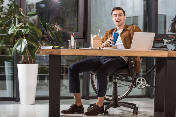 красивый счастливый бизнесмен обедает на рабочем месте в офисе и смотрит в камеру
 - Фото, изображение