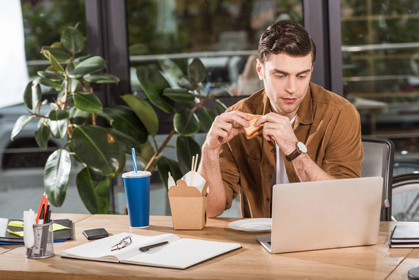 bell'uomo d'affari che mangia sandwich e toglie tagliatelle in ufficio mentre lavora con il computer portatile
 - Foto, immagini