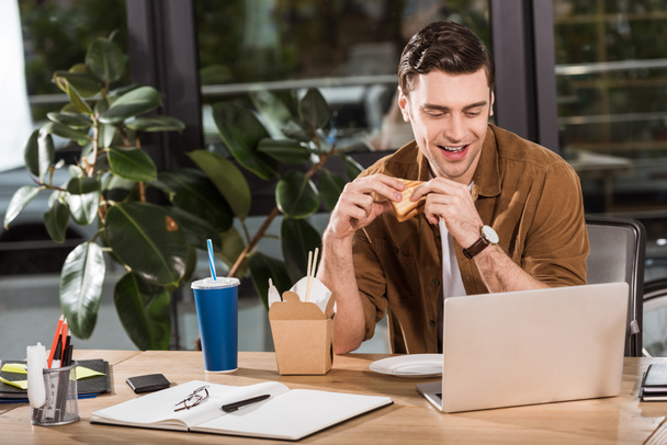 komea onnellinen liikemies syö voileipää ja ottaa pois nuudelit toimistossa työskennellessään kannettavan tietokoneen kanssa
 - Valokuva, kuva