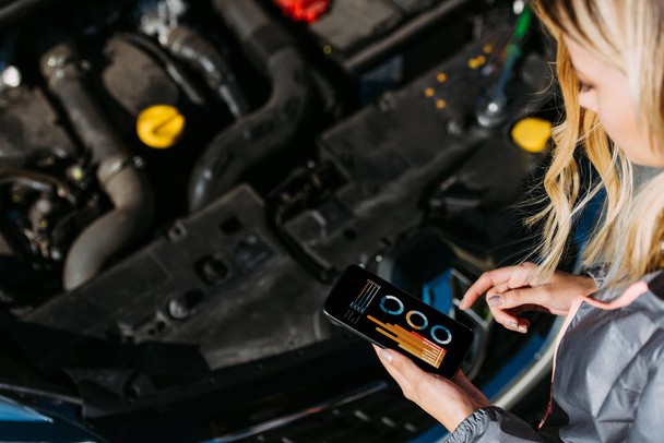cropped shot of woman using smartphone with graphs on screen while repairing broken car - Foto, Imagem