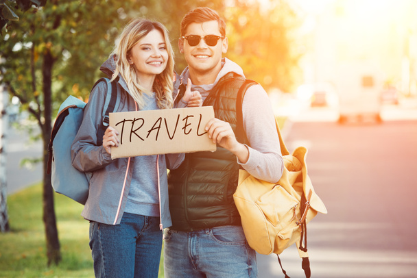 happy young couple of travelers holding card with inscription travel and smiling at camera on road - Foto, Imagen