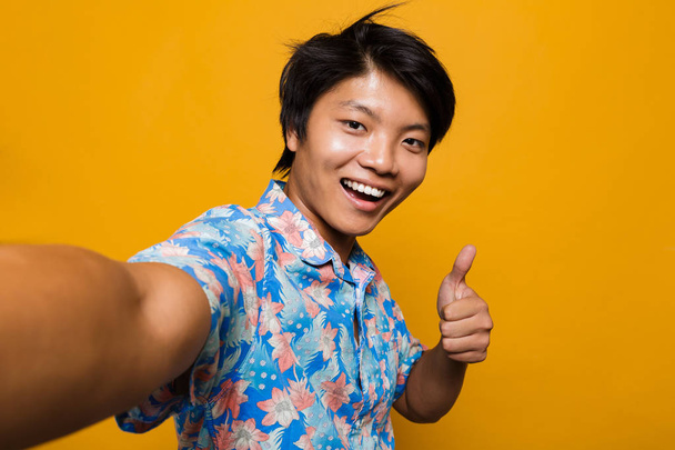 Image de jeune homme asiatique heureux posant isolé sur fond jaune prendre un selfie avec les pouces vers le haut geste
. - Photo, image