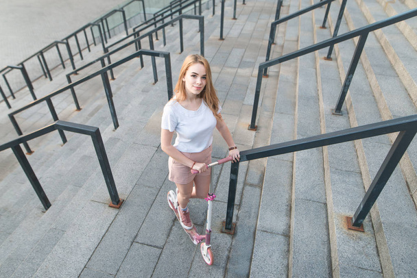 Pretty young girl with a pink kick scooter stands on the background of a modern architecture, view of the camera, top view. Copyspace - 写真・画像