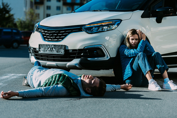 scared young woman sitting near car and looking at injured man lying on road after traffic collision - Fotoğraf, Görsel