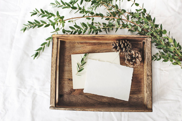 Christmas or winter wedding mock-up scene. Blank cotton paper greeting cards, old wooden tray, pine cones and green Eucalyptus parvifolia branch.White bed linen background. Flat lay, top view. - 写真・画像