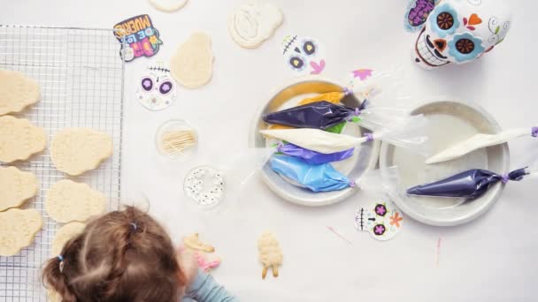 Bovenaanzicht van handen van moeder en dochter versieren van suiker schedel koekjes met royal icing voor Dia de los Muertos vakantie. - Video
