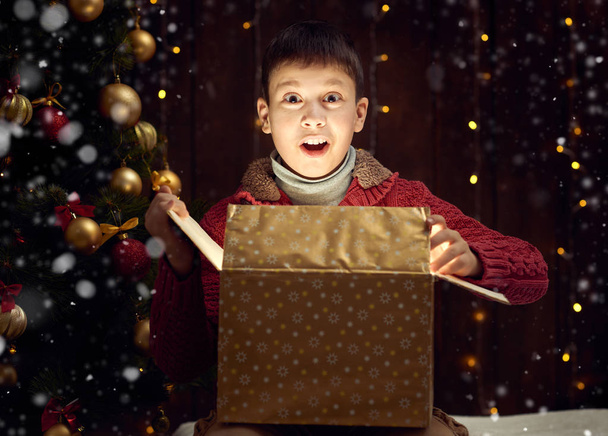 niño sentado con caja de regalo cerca de abeto decorado navidad, fondo de madera oscura
 - Foto, Imagen
