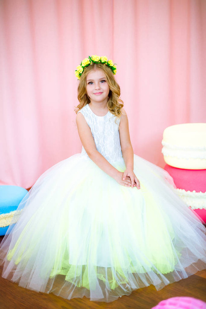 Cute little girl with blond curls in a white lush dress with giant sweets in pink background studio - Foto, Imagen