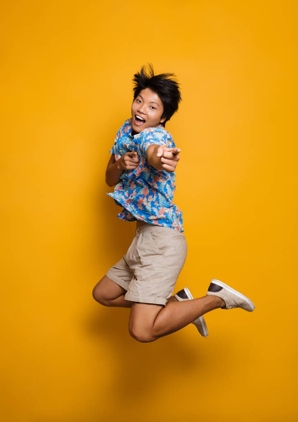 Image of happy emotional young asian man jumping isolated over yellow background pointing. - Valokuva, kuva