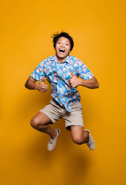 Image of happy emotional young asian man jumping isolated over yellow background showing thumbs up. - Fotó, kép
