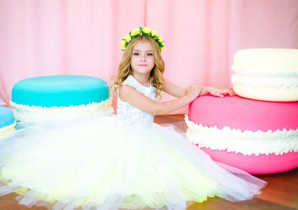 Cute little girl with blond curls in a white lush dress with giant sweets in pink background studio - Foto, imagen