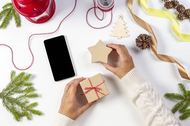 Kerst schaven, online winkelen concept. De handen van de vrouw met de mobiele telefoon, kerstcadeautjes, decoraties op witte achtergrond - Foto, afbeelding