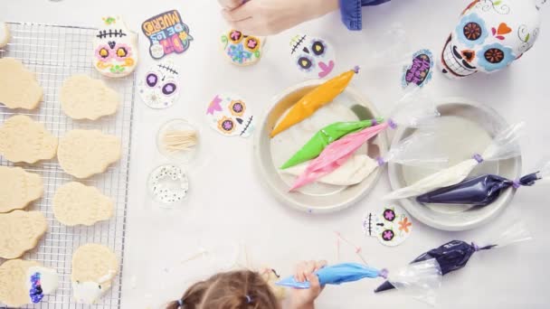 Bovenaanzicht van handen van moeder en dochter versieren van suiker schedel koekjes met royal icing voor Dia de los Muertos vakantie. - Video