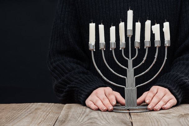 cropped shot of woman standing at wooden tabletop with menorah and candles on black background, hannukah celebration concept - Fotó, kép
