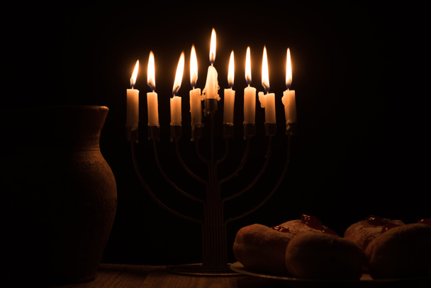 close up view of traditional sweet doughnuts, clay jug and menorah with candles on black background, hannukah holiday concept - Photo, Image