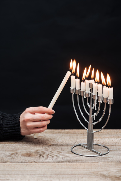 partial view of woman lighting candles on menorah on wooden tabletop on black backdrop, hannukah holiday concept - Photo, Image