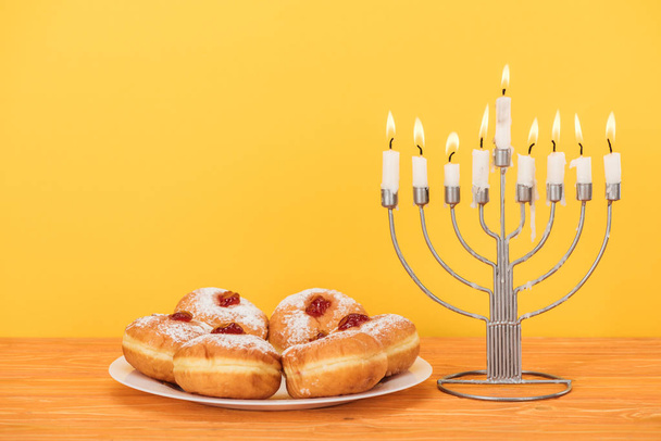 Nahaufnahme von süßen Donuts und Menora mit Kerzen auf hölzerner Oberfläche isoliert auf gelbem, hannukah Konzept - Foto, Bild